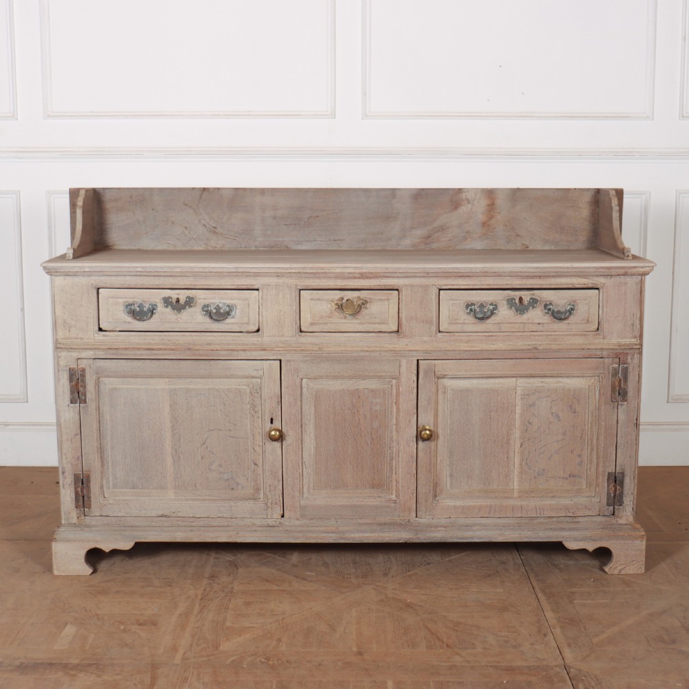 An 18th-century bleached oak dresser base featuring three top drawers, panelled cupboard doors, and a simple low wooden gallery back.