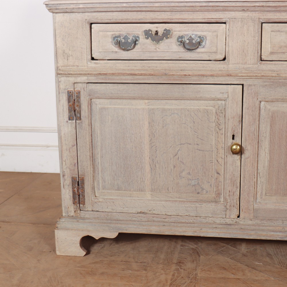 An 18th-century bleached oak dresser base featuring three top drawers, panelled cupboard doors, and a simple low wooden gallery back. View IV