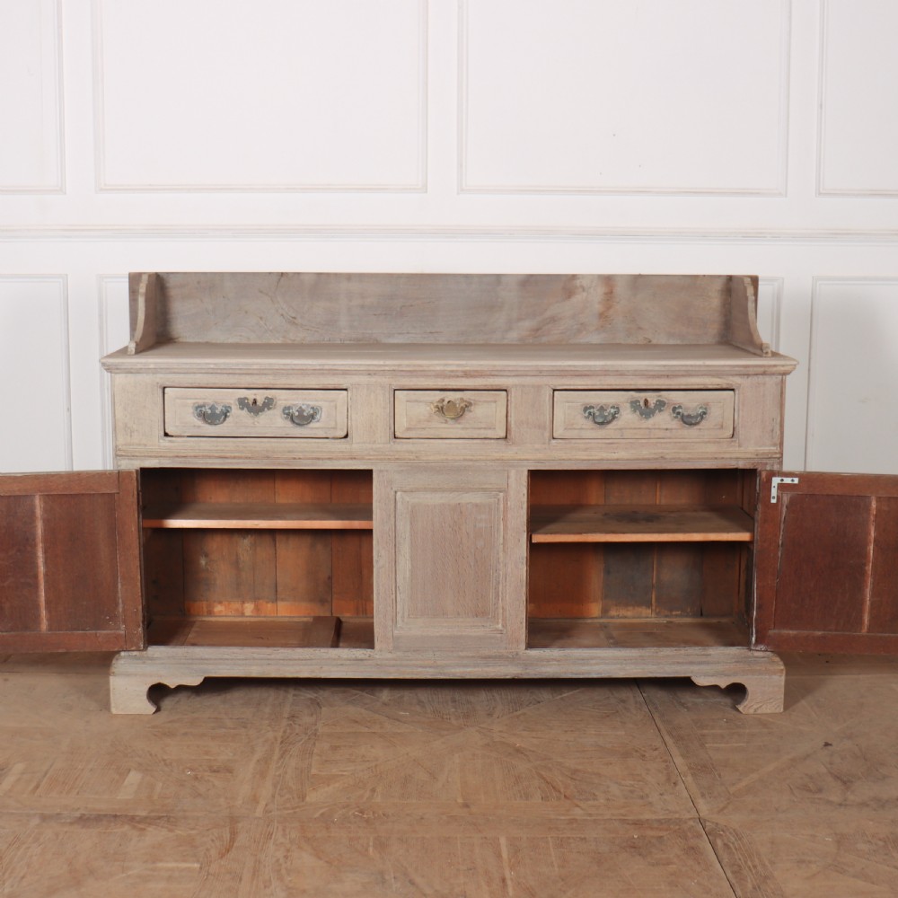 An 18th-century bleached oak dresser base featuring three top drawers, panelled cupboard doors, and a simple low wooden gallery back. View V