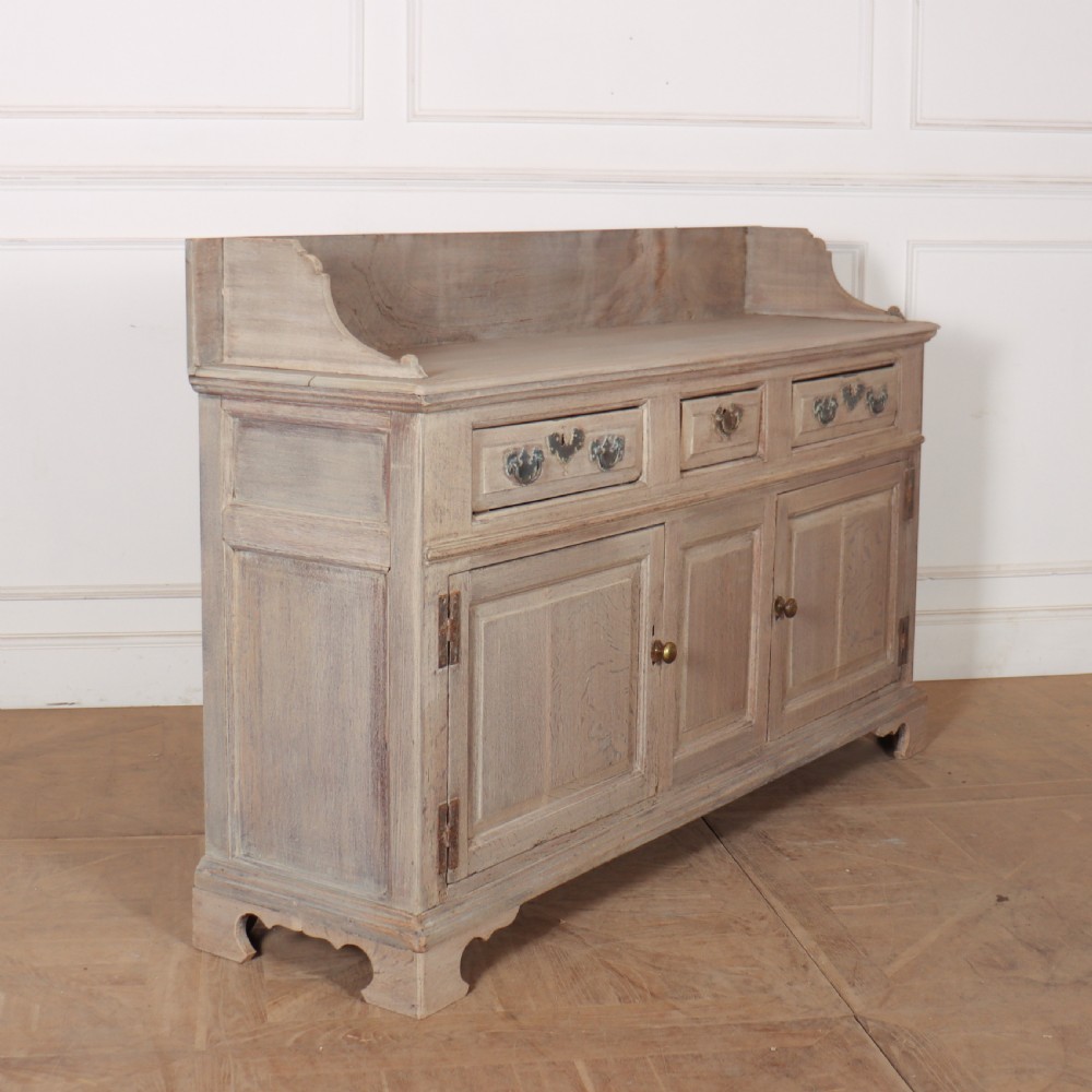 An 18th-century bleached oak dresser base featuring three top drawers, panelled cupboard doors, and a simple low wooden gallery back. View VI