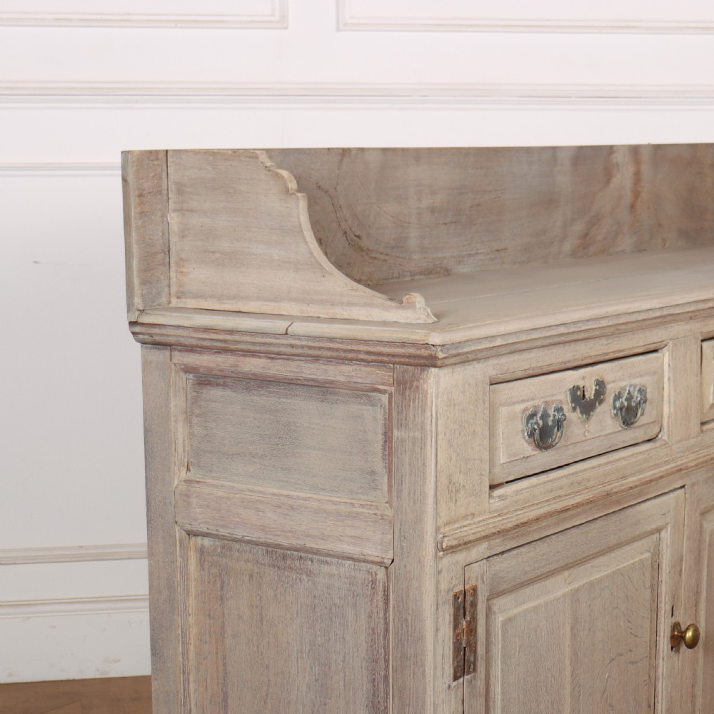 An 18th-century bleached oak dresser base featuring three top drawers, panelled cupboard doors, and a simple low wooden gallery back. View VII