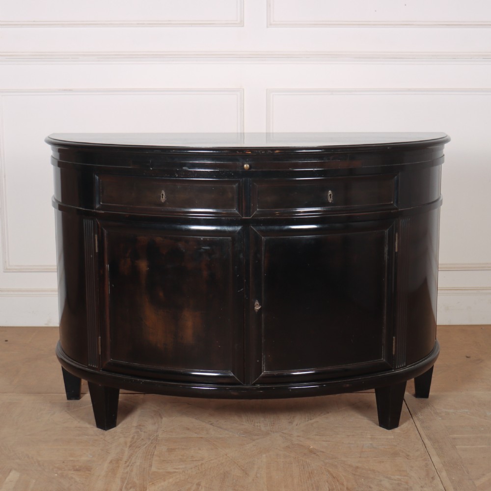 A French ebonised demi lune cabinet featuring a semi-circular body, top drawers with keyhole escutcheons, panelled cupboard doors, and square tapered feet.