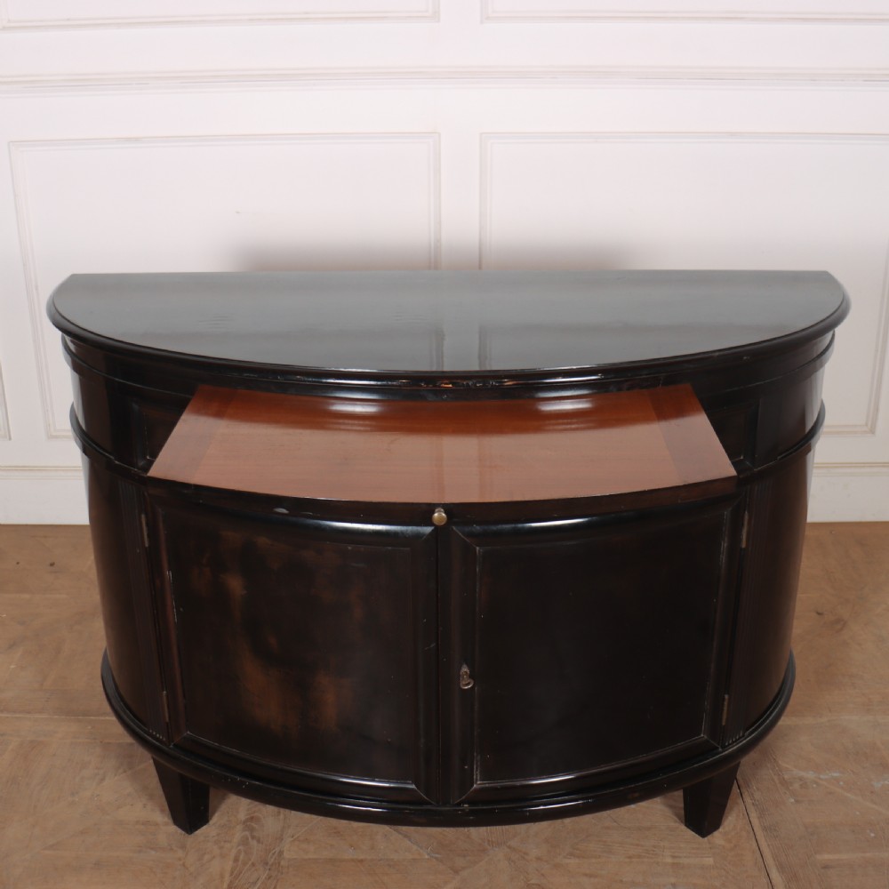 A French ebonised demi lune cabinet featuring a semi-circular body, top drawers with keyhole escutcheons, panelled cupboard doors, and square tapered feet. View II