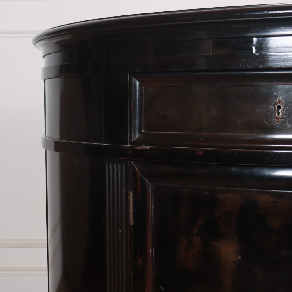 A French ebonised demi lune cabinet featuring a semi-circular body, top drawers with keyhole escutcheons, panelled cupboard doors, and square tapered feet. View IV