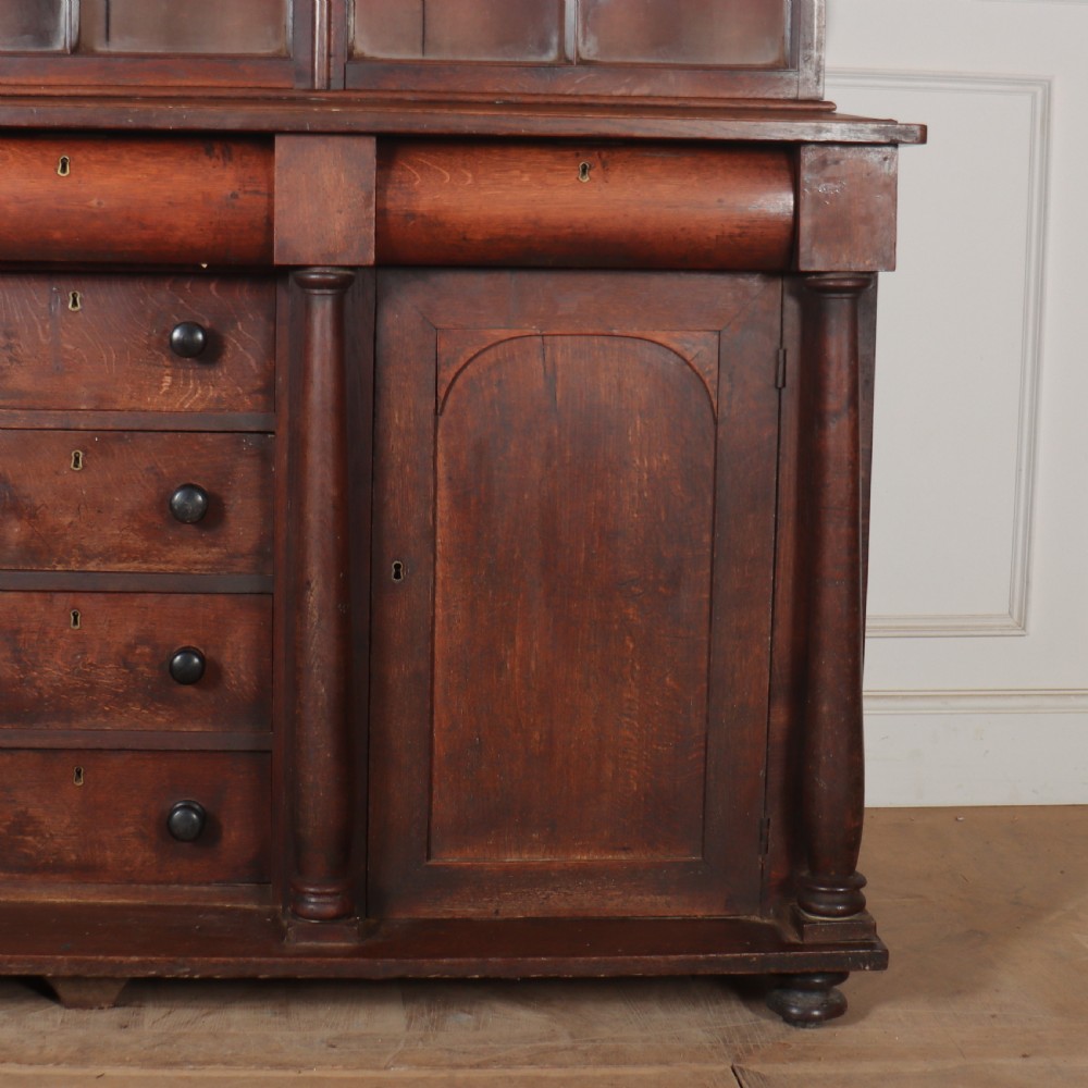 A dark brown antique oak glazed dresser featuring a upper display cabinet with three glass-front doors above a base with drawers and cupboard doors. View V