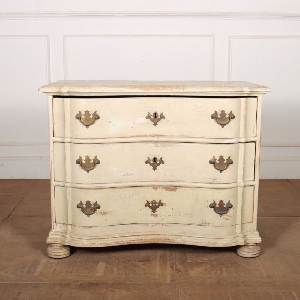 An 18th-century serpentine commode featuring three drawers with a distressed cream paint finish and cast brass hardware.