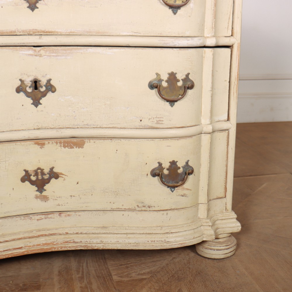An 18th-century serpentine commode featuring three drawers with a distressed cream paint finish and cast brass hardware. View V