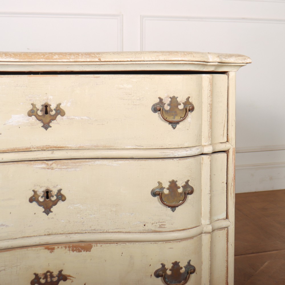 An 18th-century serpentine commode featuring three drawers with a distressed cream paint finish and cast brass hardware. View VI