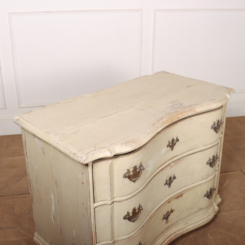 An 18th-century serpentine commode featuring three drawers with a distressed cream paint finish and cast brass hardware. View VII