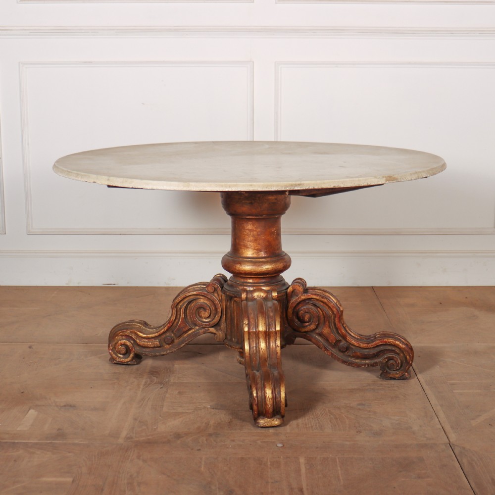 An antique circular table featuring a cream marble top resting upon a heavily carved giltwood pedestal base with four scroll feet.