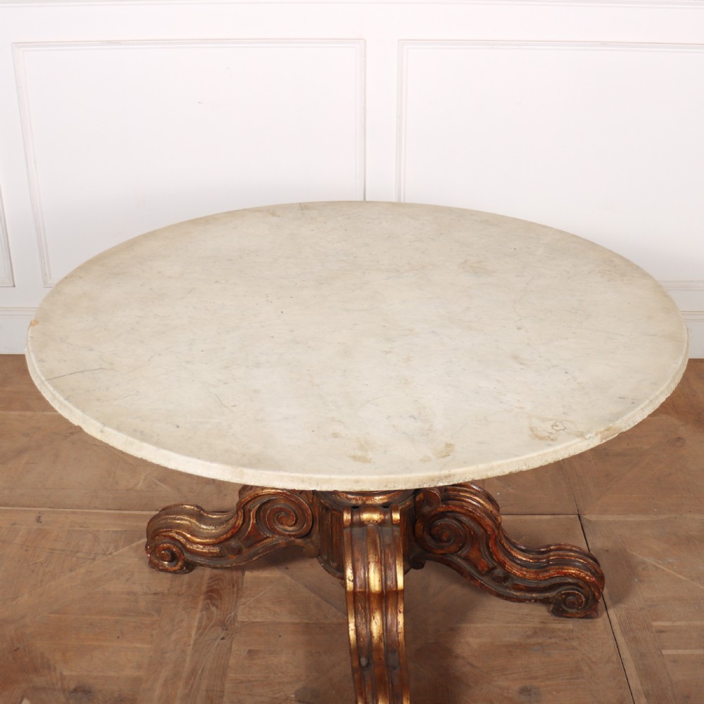 An antique circular table featuring a cream marble top resting upon a heavily carved giltwood pedestal base with four scroll feet. View IV