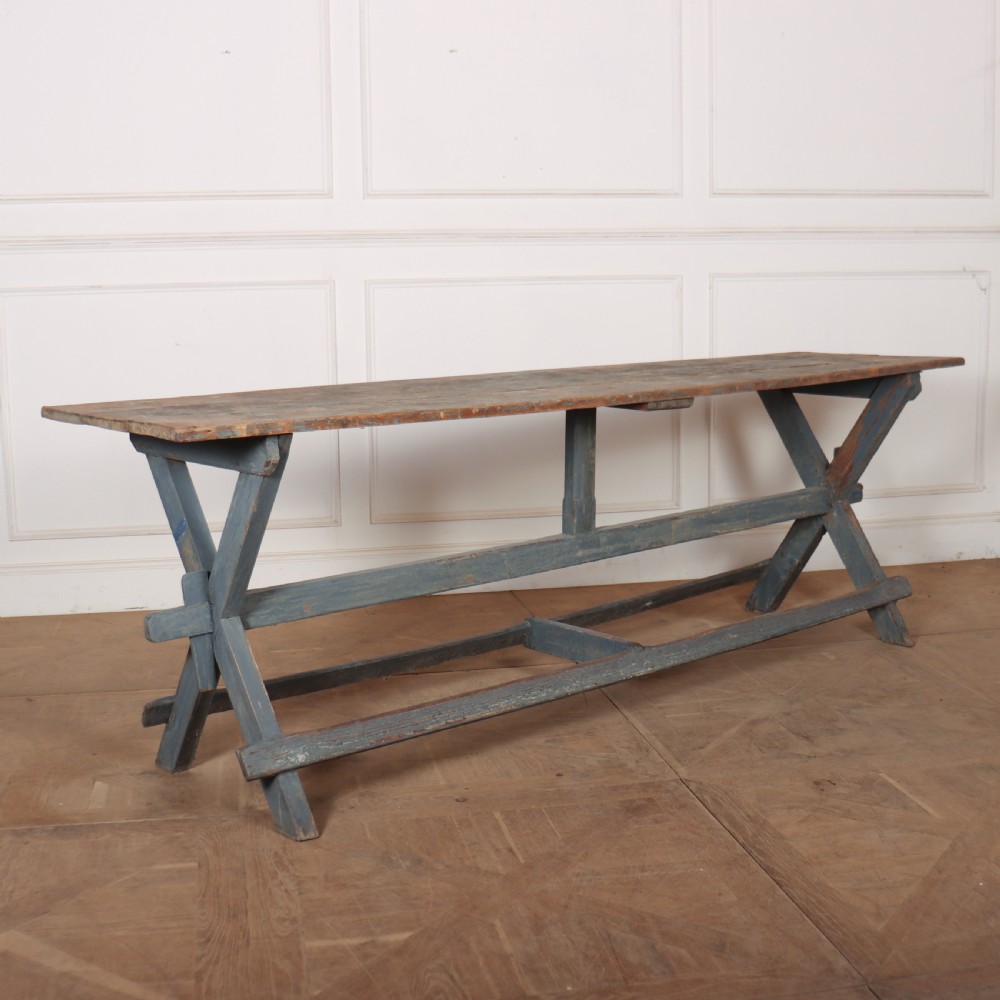 A long antique wooden tavern table with a natural wood plank top and a blue-painted X-frame trestle base.