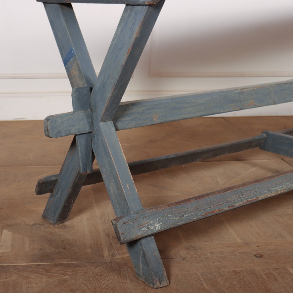 A long antique wooden tavern table with a natural wood plank top and a blue-painted X-frame trestle base. View III