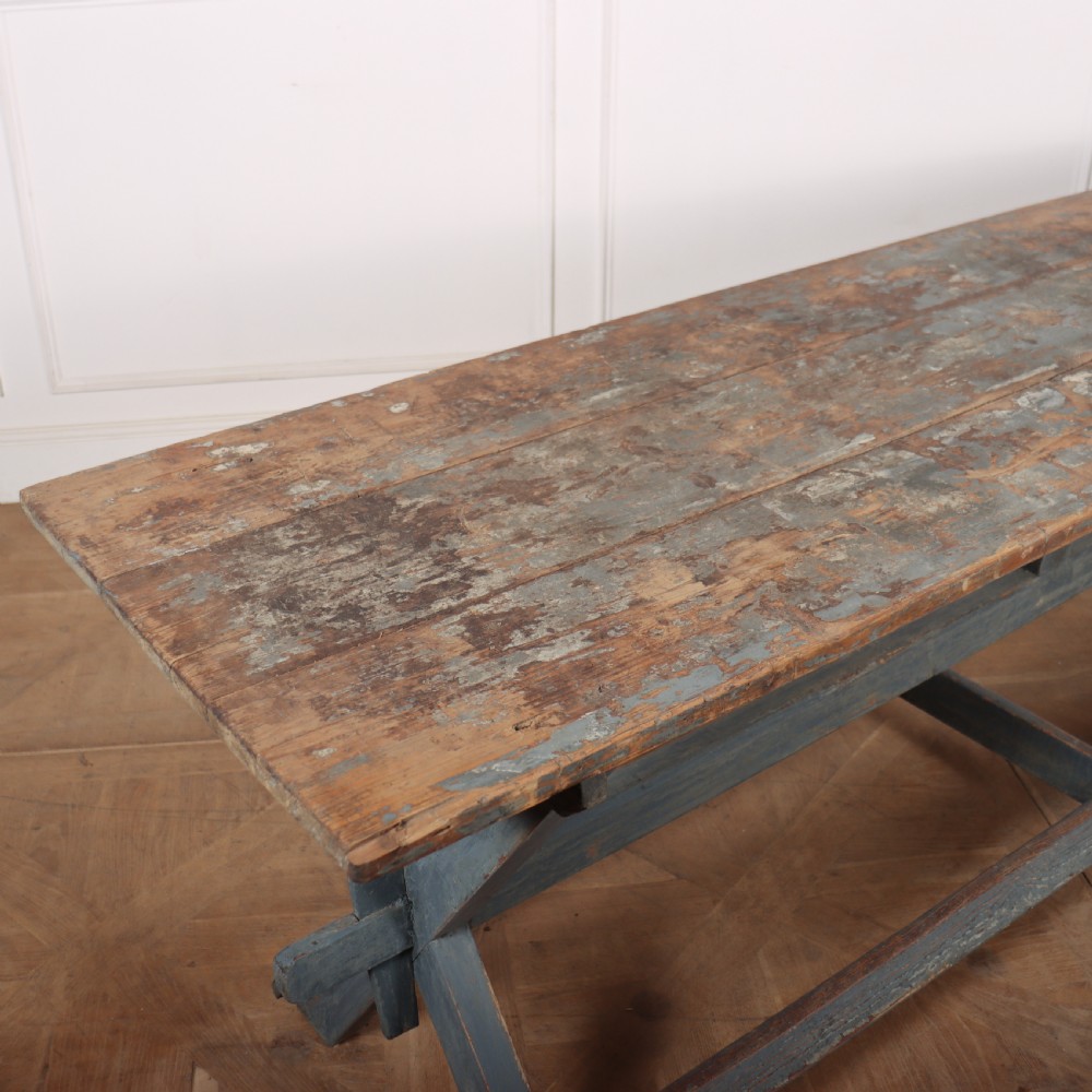 A long antique wooden tavern table with a natural wood plank top and a blue-painted X-frame trestle base. View V