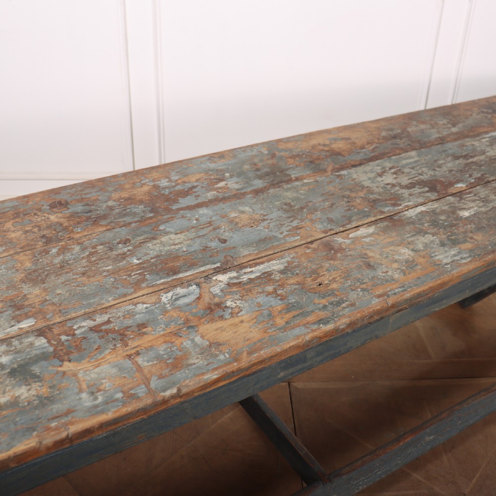 A long antique wooden tavern table with a natural wood plank top and a blue-painted X-frame trestle base. View VI