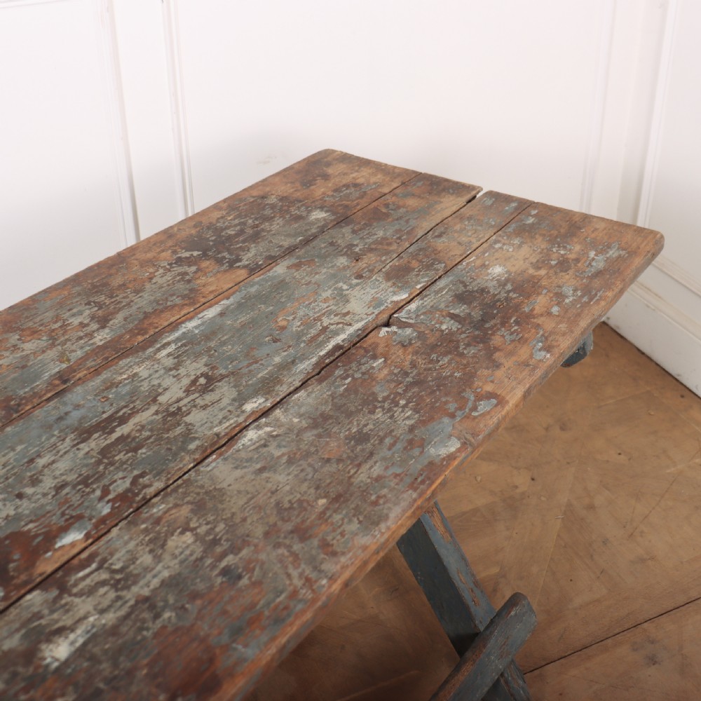 A long antique wooden tavern table with a natural wood plank top and a blue-painted X-frame trestle base. View VII