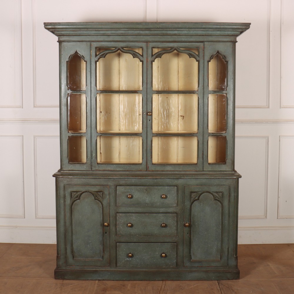 A two-part blue-green painted timber dresser featuring an upper glazed cabinet with gothic arch doors and a lower base fitted with drawers and cupboard doors.