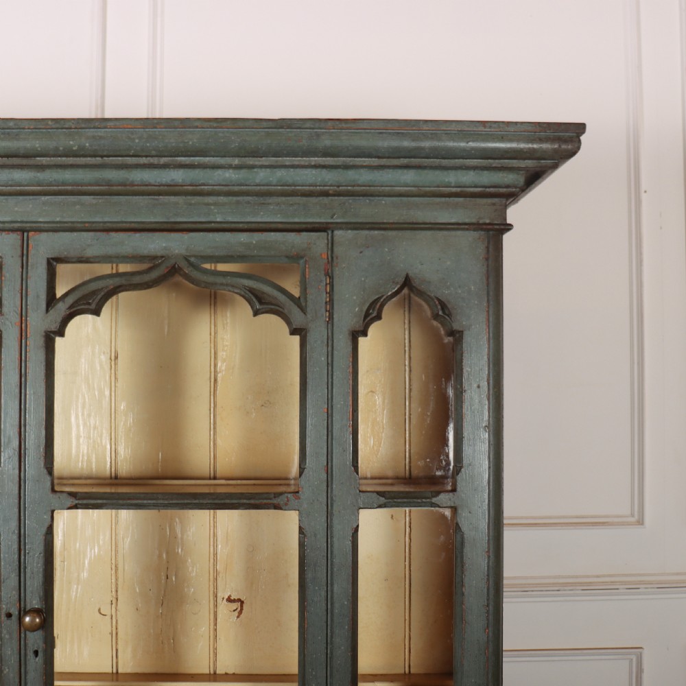 A two-part blue-green painted timber dresser featuring an upper glazed cabinet with gothic arch doors and a lower base fitted with drawers and cupboard doors. View VI