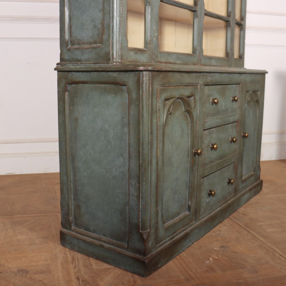 A two-part blue-green painted timber dresser featuring an upper glazed cabinet with gothic arch doors and a lower base fitted with drawers and cupboard doors. View X