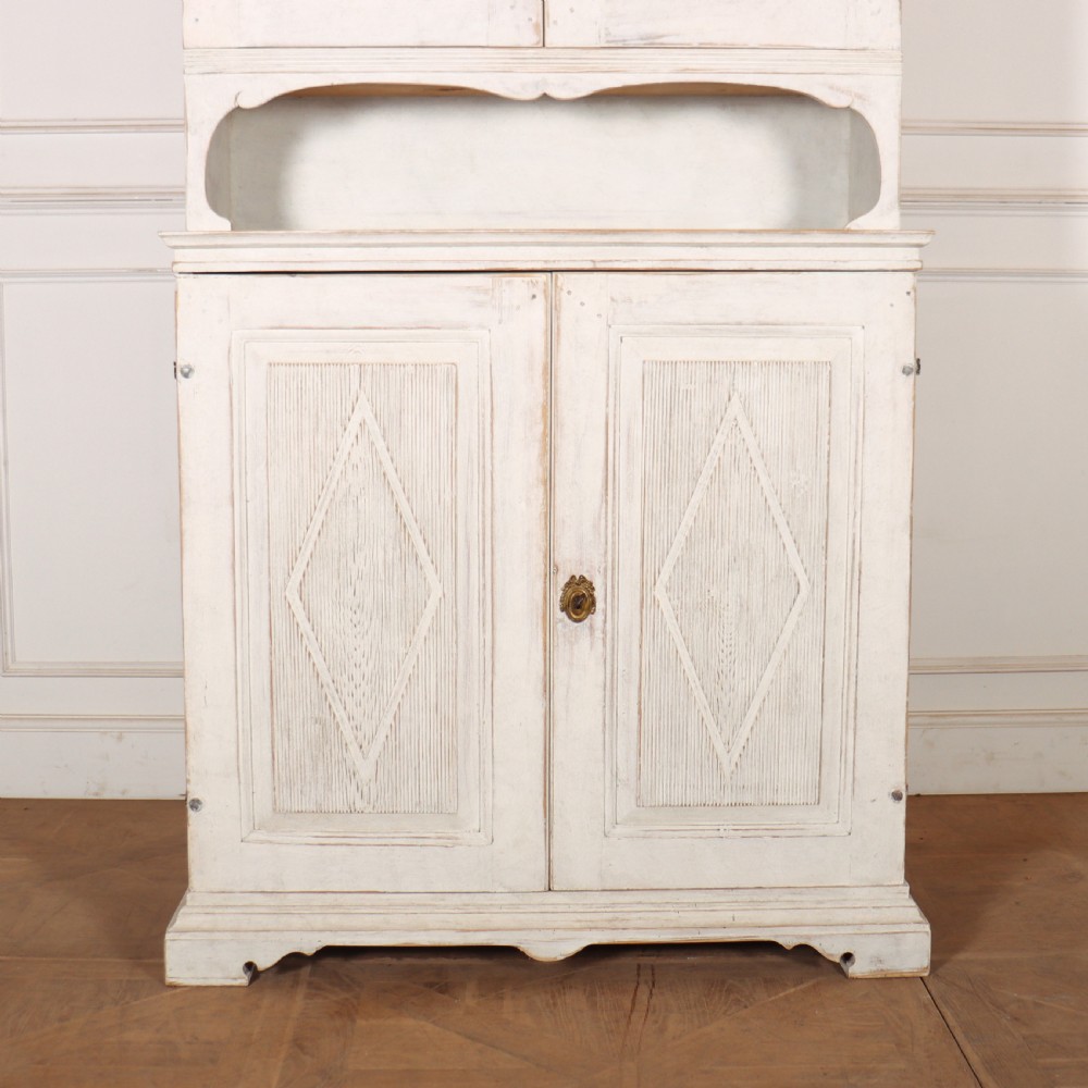 A two-tier Swedish painted wooden cabinet featuring distressed off-white paint, carved diamond geometric panels on four doors, and central brass escutcheons. View VI