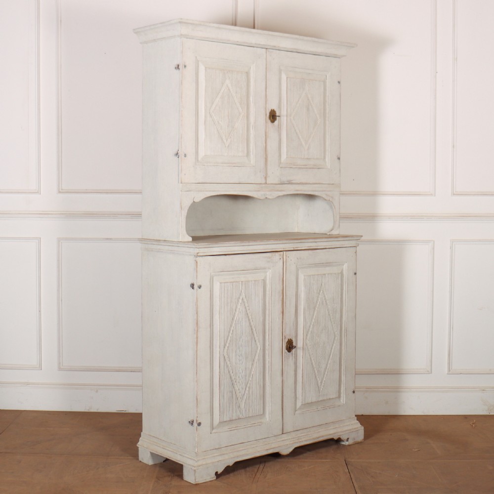 A two-tier Swedish painted wooden cabinet featuring distressed off-white paint, carved diamond geometric panels on four doors, and central brass escutcheons. View VII