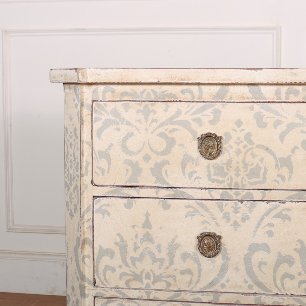 A Swedish painted pine commode featuring three drawers decorated with off-white and grey damask pattern paintwork, brass escutcheons, and tapered square legs. View II