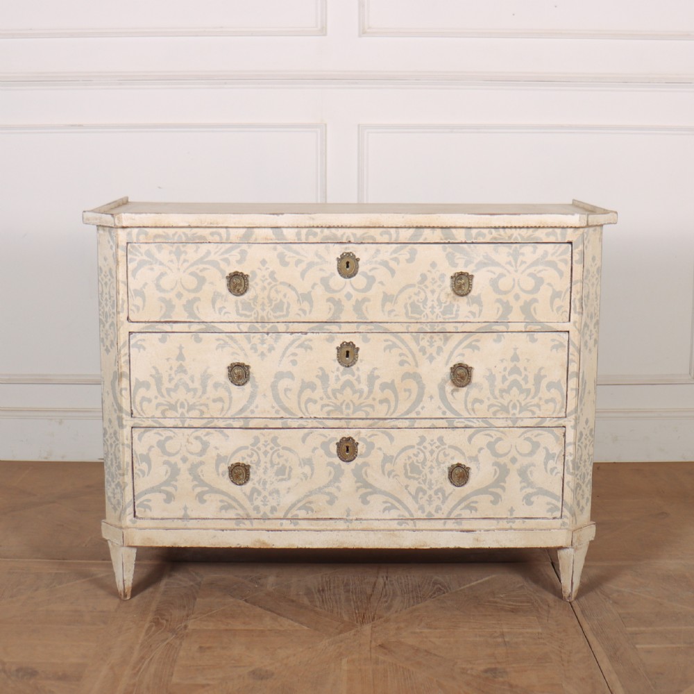 A Swedish painted pine commode featuring three drawers decorated with off-white and grey damask pattern paintwork, brass escutcheons, and tapered square legs.