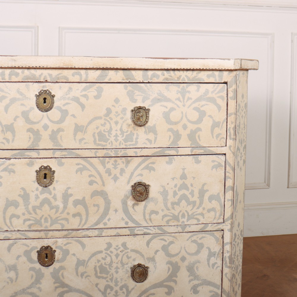 A Swedish painted pine commode featuring three drawers decorated with off-white and grey damask pattern paintwork, brass escutcheons, and tapered square legs. View V