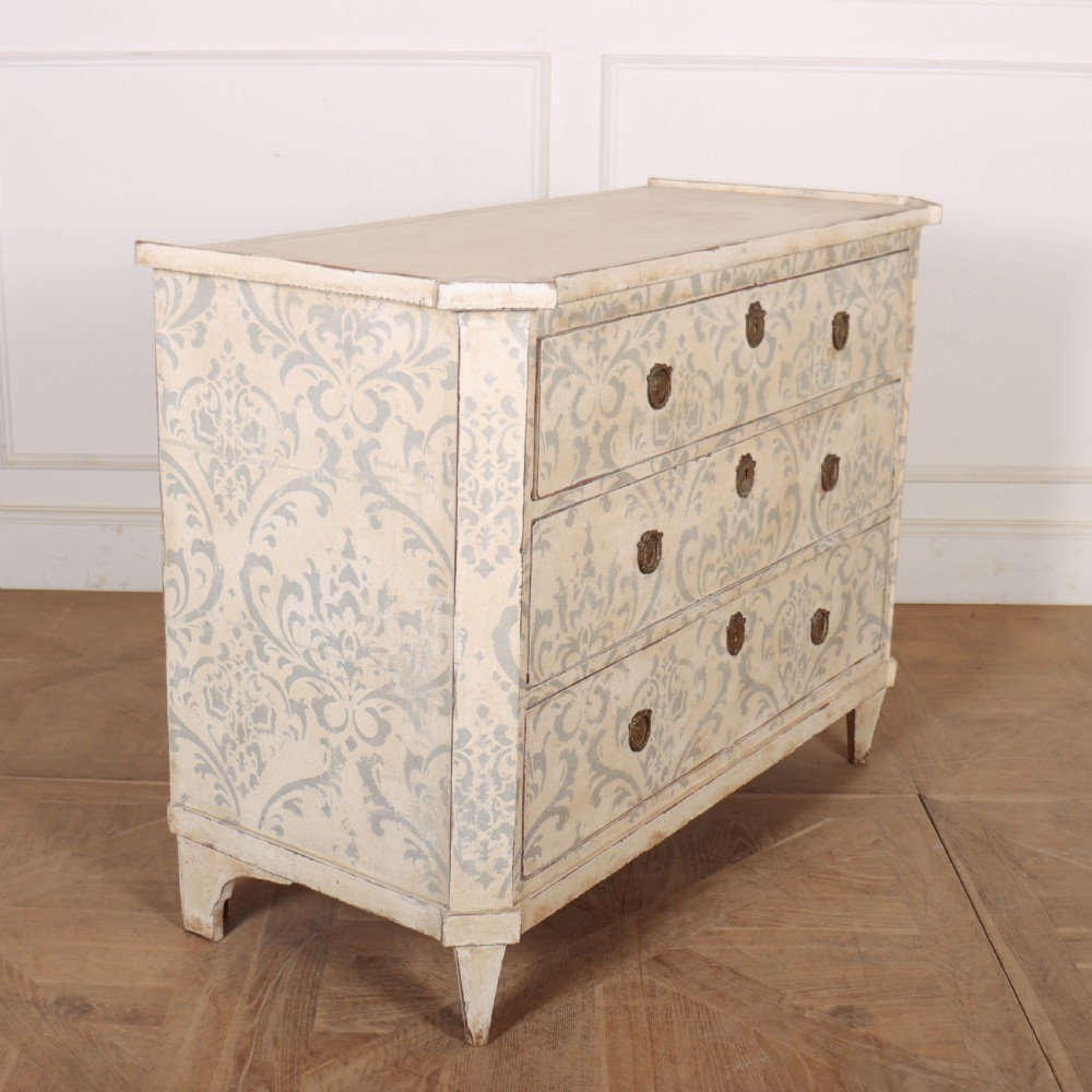 A Swedish painted pine commode featuring three drawers decorated with off-white and grey damask pattern paintwork, brass escutcheons, and tapered square legs. View VIII