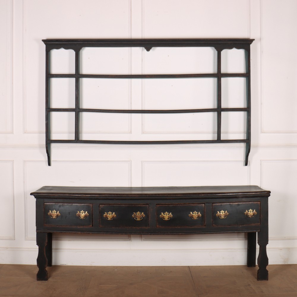 An 18th-century dark painted wooden kitchen dresser base featuring four drawers with brass handles beneath an open wall-mounted plate rack.