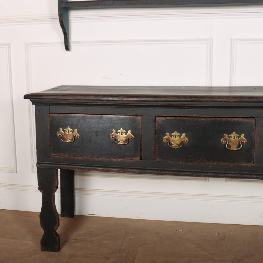An 18th-century dark painted wooden kitchen dresser base featuring four drawers with brass handles beneath an open wall-mounted plate rack. View II