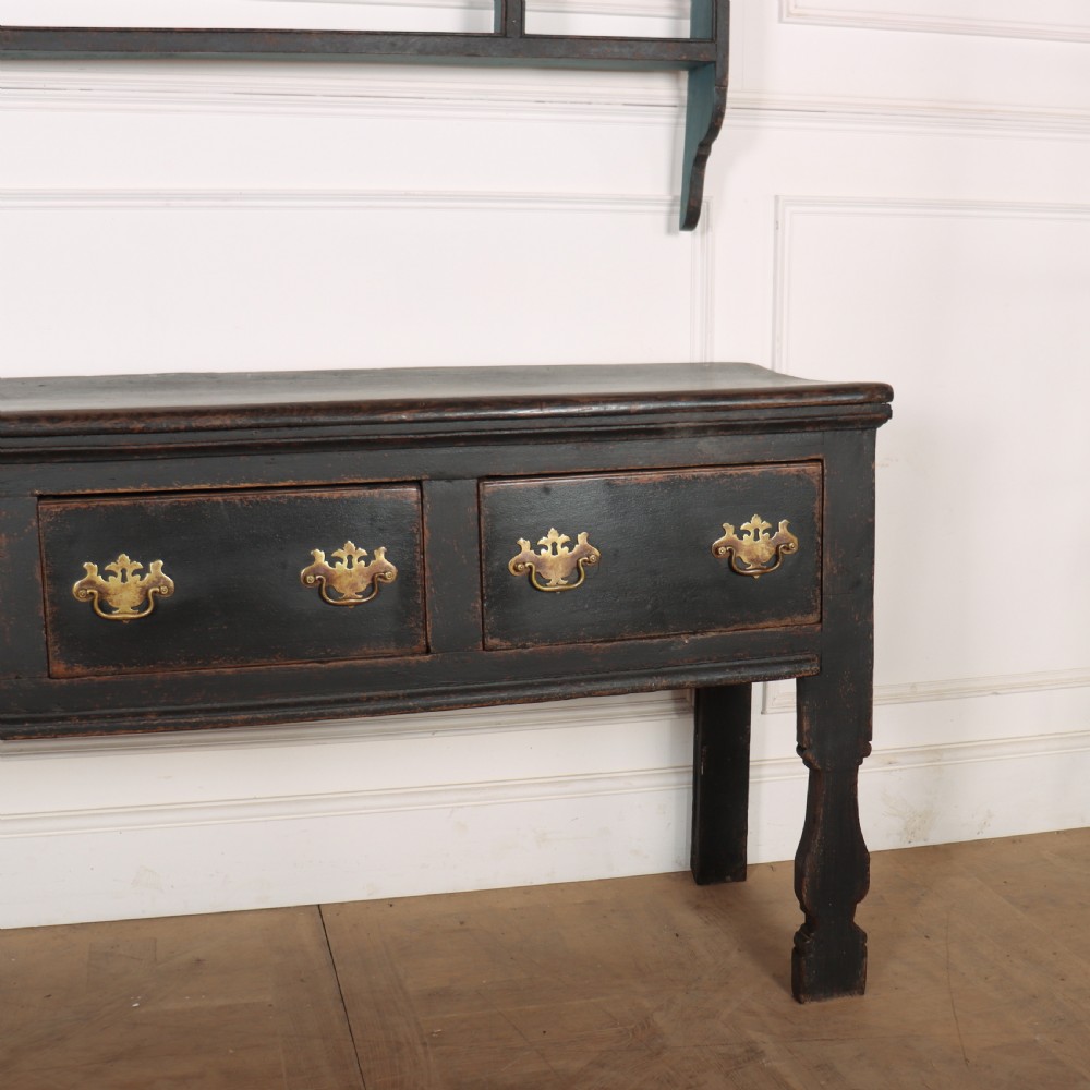 An 18th-century dark painted wooden kitchen dresser base featuring four drawers with brass handles beneath an open wall-mounted plate rack. View III