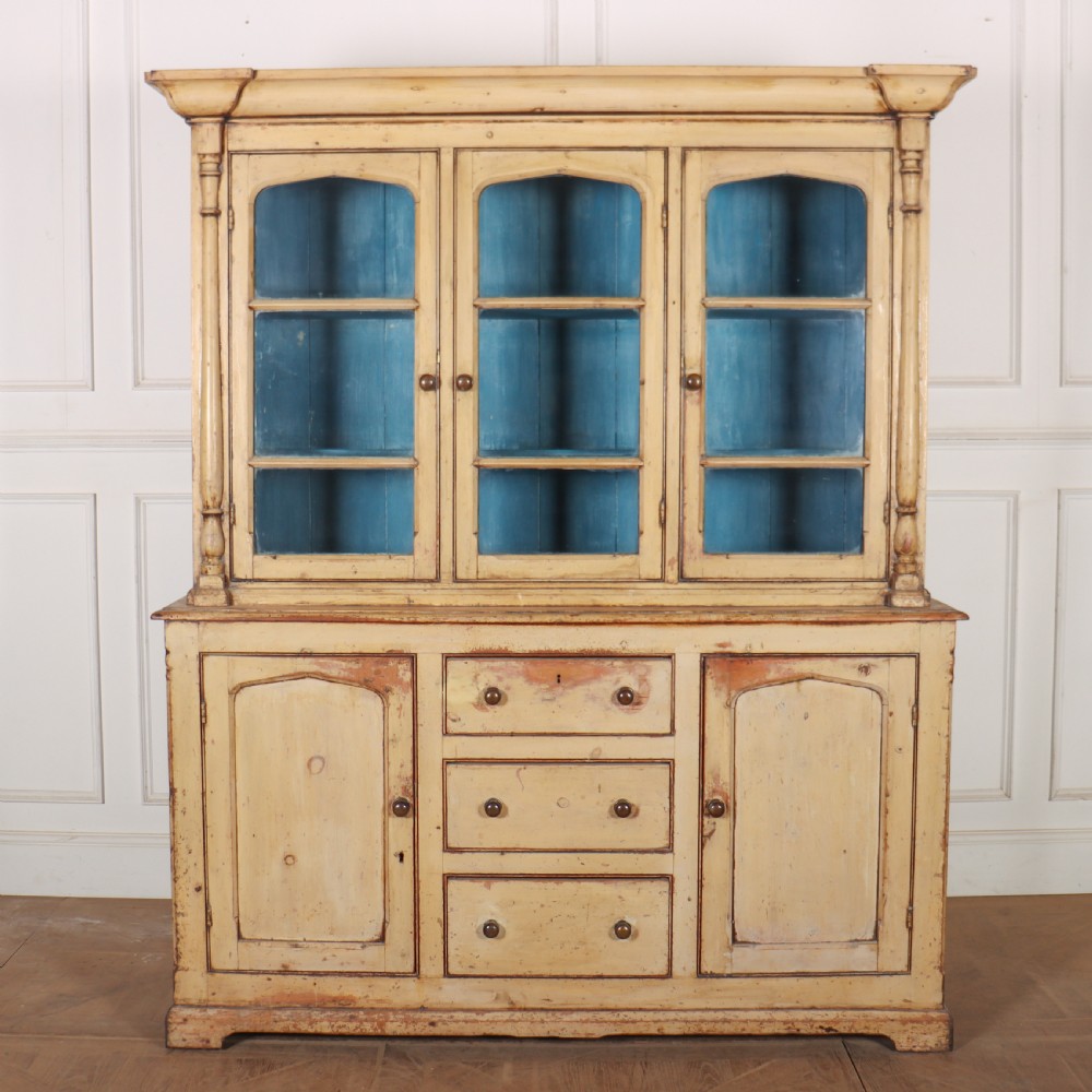A cream painted timber kitchen dresser featuring a glazed three-door upper display cabinet with a vibrant blue interior above a base with drawers and panelled cupboard doors.