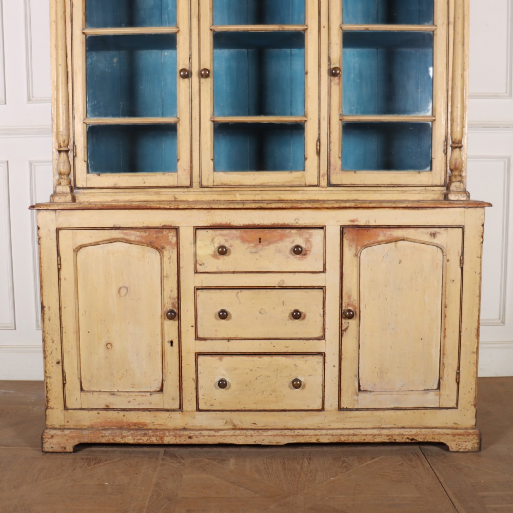 A cream painted timber kitchen dresser featuring a glazed three-door upper display cabinet with a vibrant blue interior above a base with drawers and panelled cupboard doors. View V