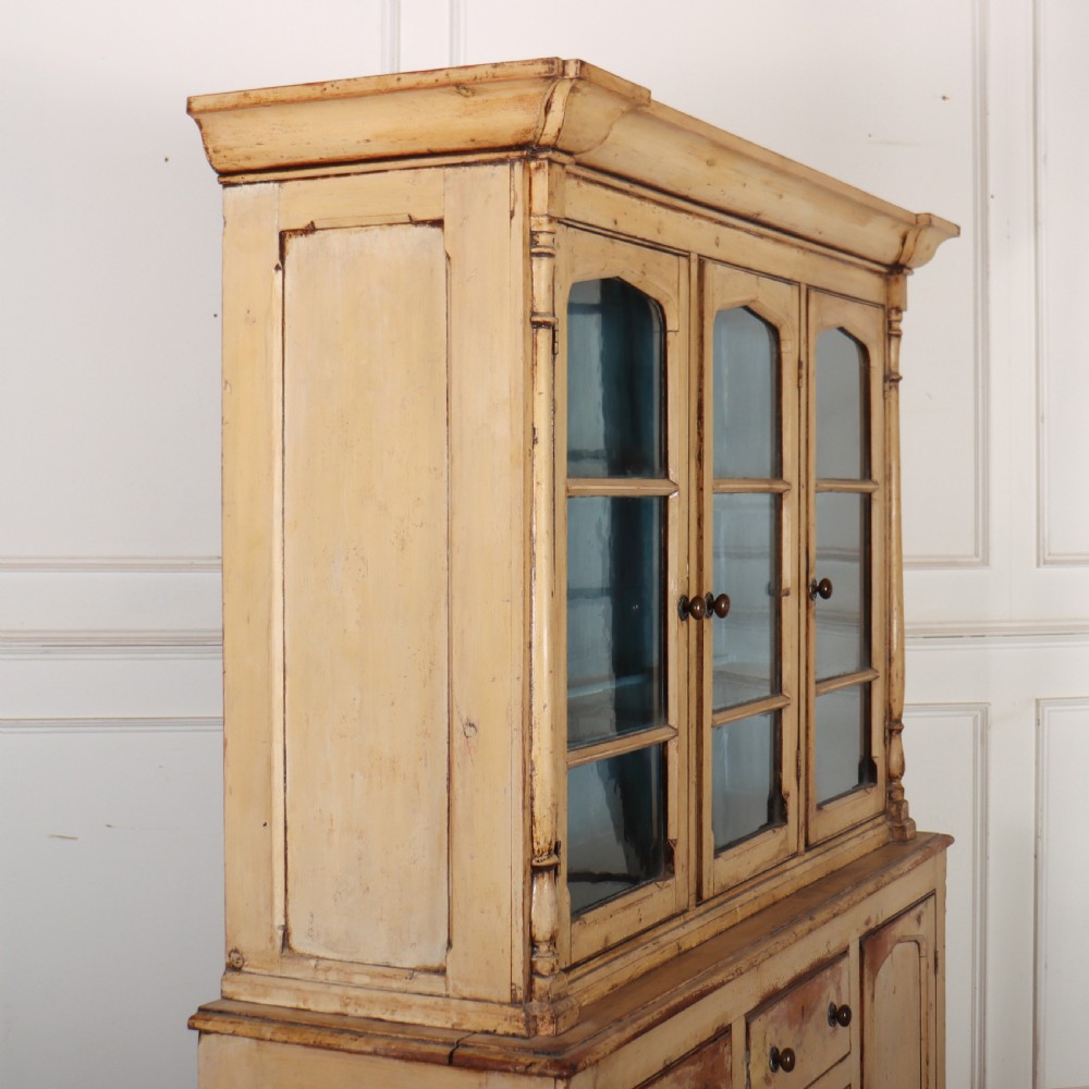 A cream painted timber kitchen dresser featuring a glazed three-door upper display cabinet with a vibrant blue interior above a base with drawers and panelled cupboard doors. View VI