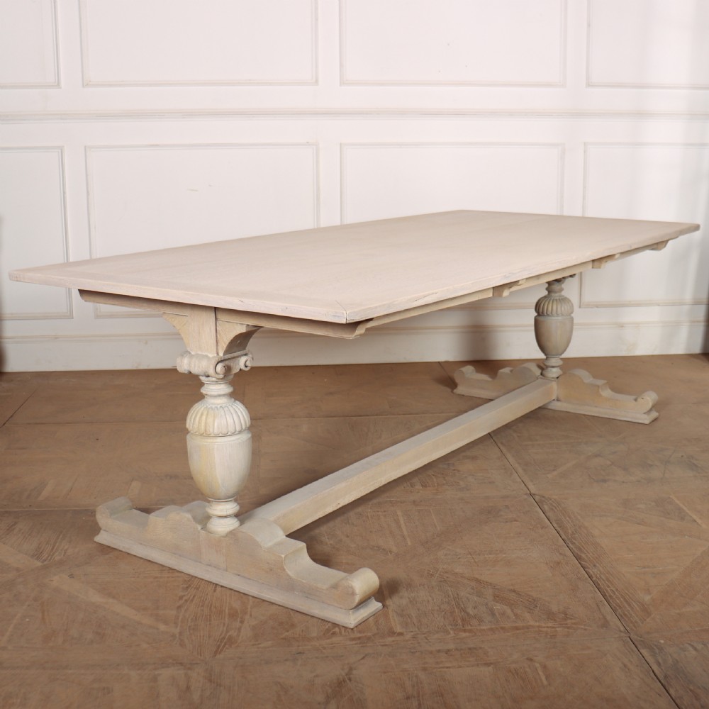 A long bleached oak dining table with a rectangular top and carved turned trestle base connected by a low central stretcher.