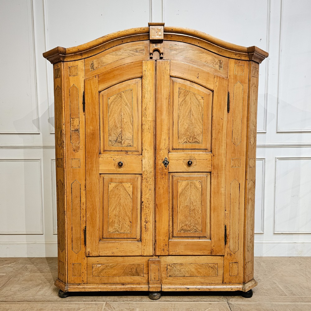 An 18th-century German pine armoire featuring an arched cornice, central cresting, geometric inlaid panel doors, and bun feet.