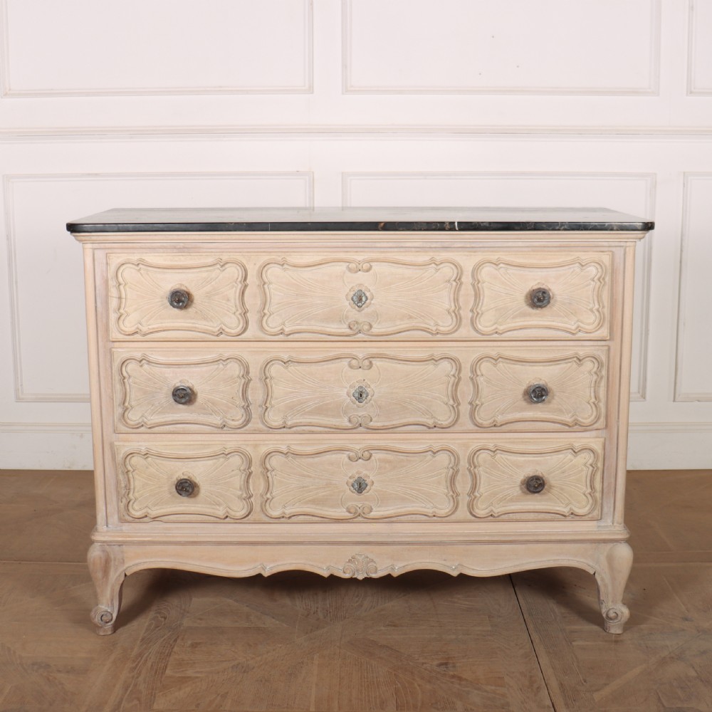A French bleached oak commode featuring three carved panel drawers with dark metal handles, keyhole escutcheons, a black marble top, and cabriole feet.
