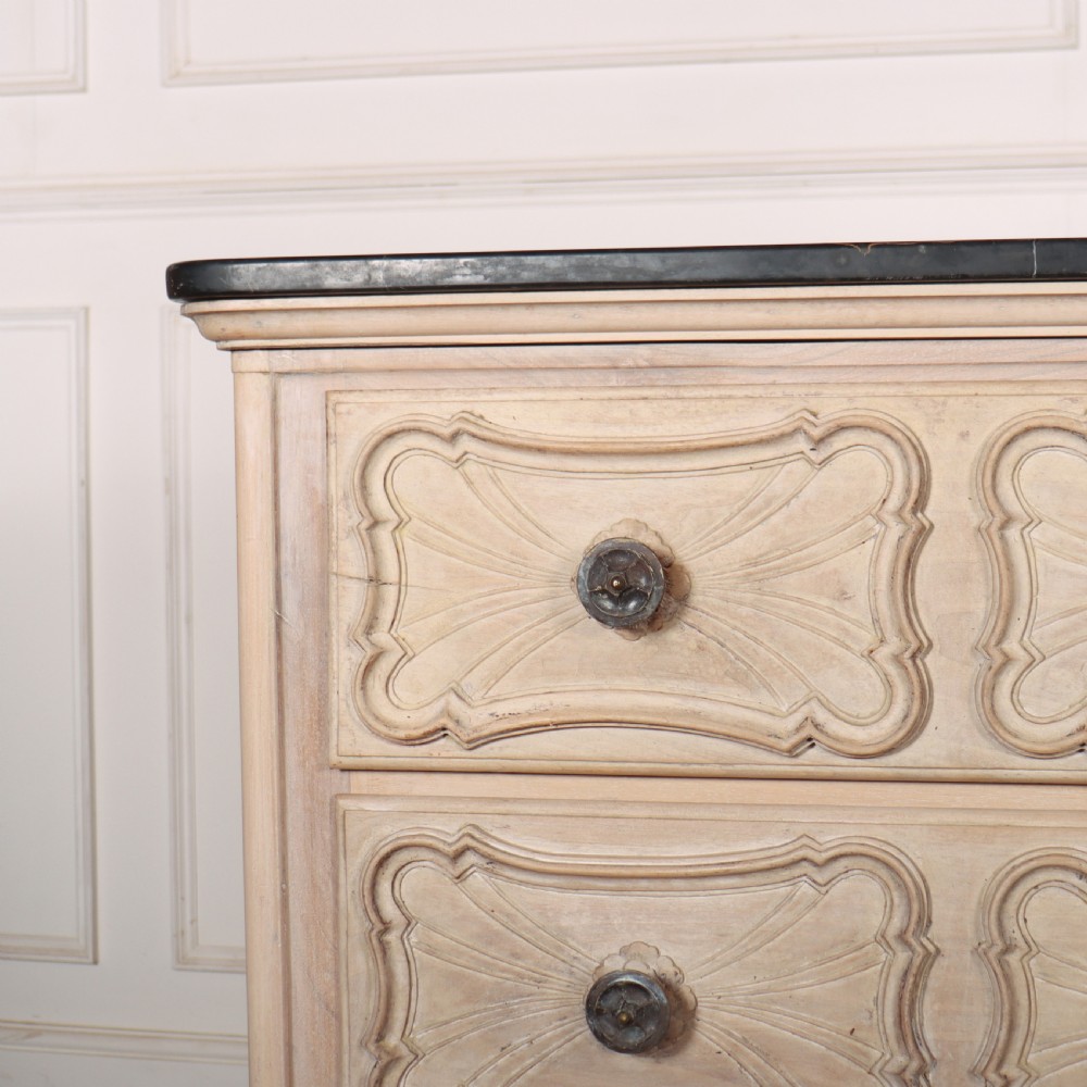 A French bleached oak commode featuring three carved panel drawers with dark metal handles, keyhole escutcheons, a black marble top, and cabriole feet. View II