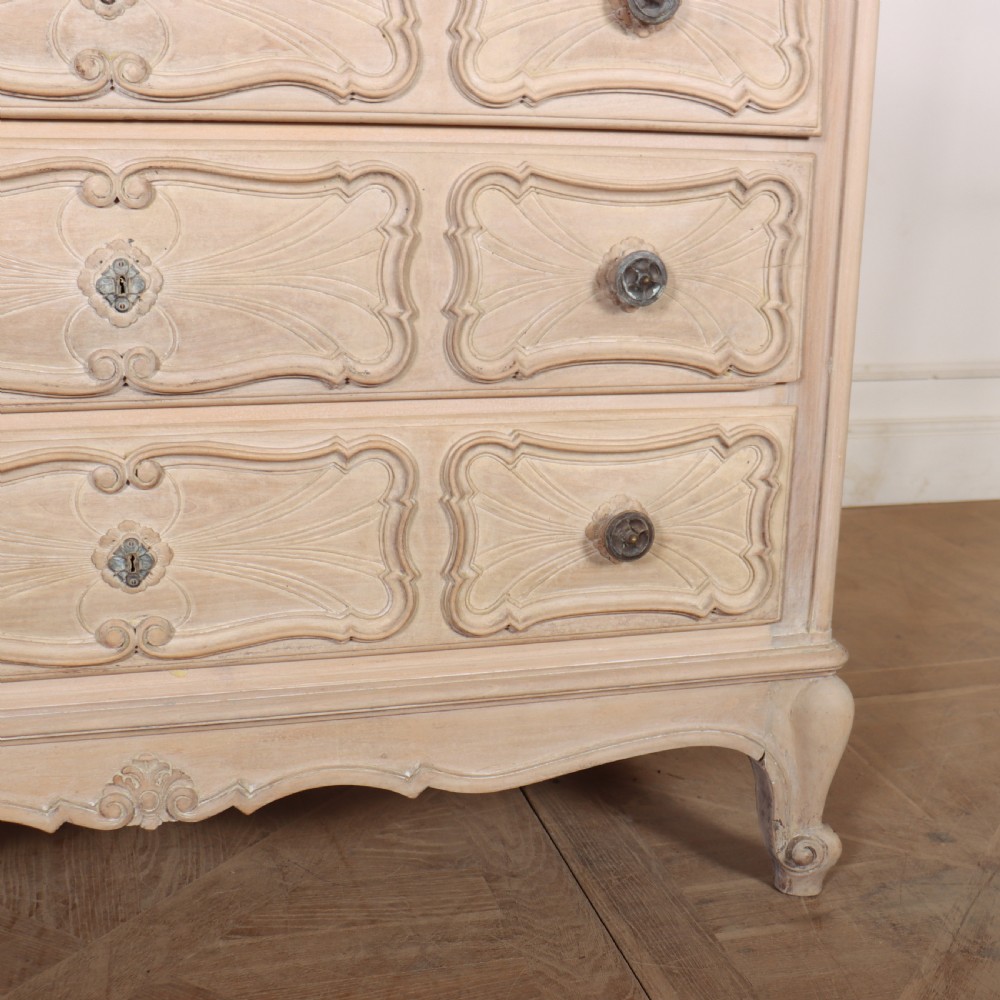 A French bleached oak commode featuring three carved panel drawers with dark metal handles, keyhole escutcheons, a black marble top, and cabriole feet. View V