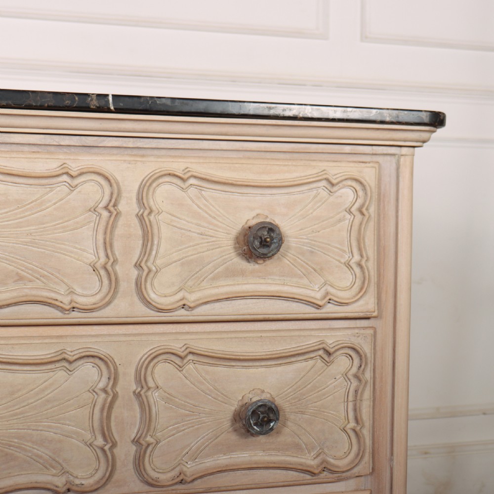 A French bleached oak commode featuring three carved panel drawers with dark metal handles, keyhole escutcheons, a black marble top, and cabriole feet. View VI
