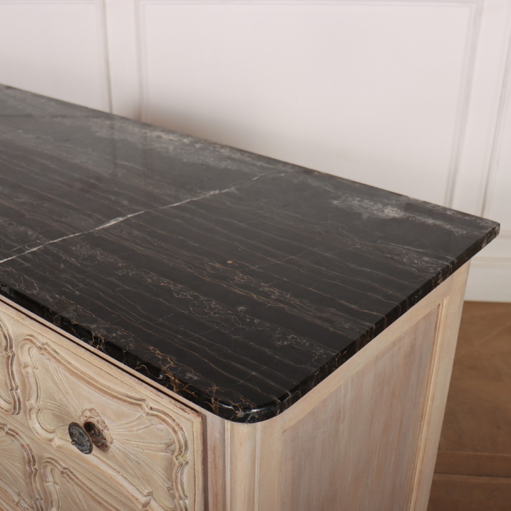 A French bleached oak commode featuring three carved panel drawers with dark metal handles, keyhole escutcheons, a black marble top, and cabriole feet. View VII
