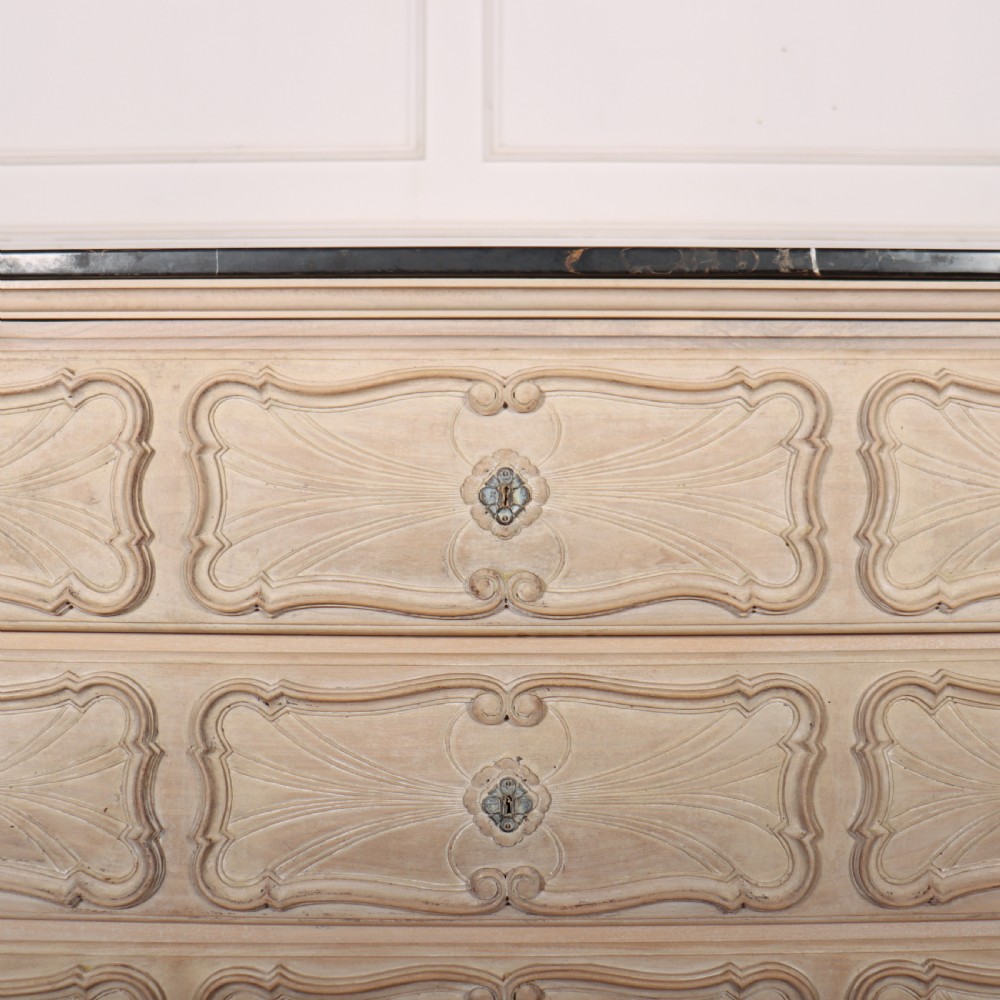 A French bleached oak commode featuring three carved panel drawers with dark metal handles, keyhole escutcheons, a black marble top, and cabriole feet. View VIII