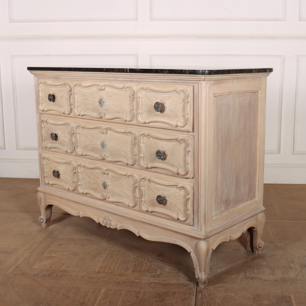 A French bleached oak commode featuring three carved panel drawers with dark metal handles, keyhole escutcheons, a black marble top, and cabriole feet. View IX