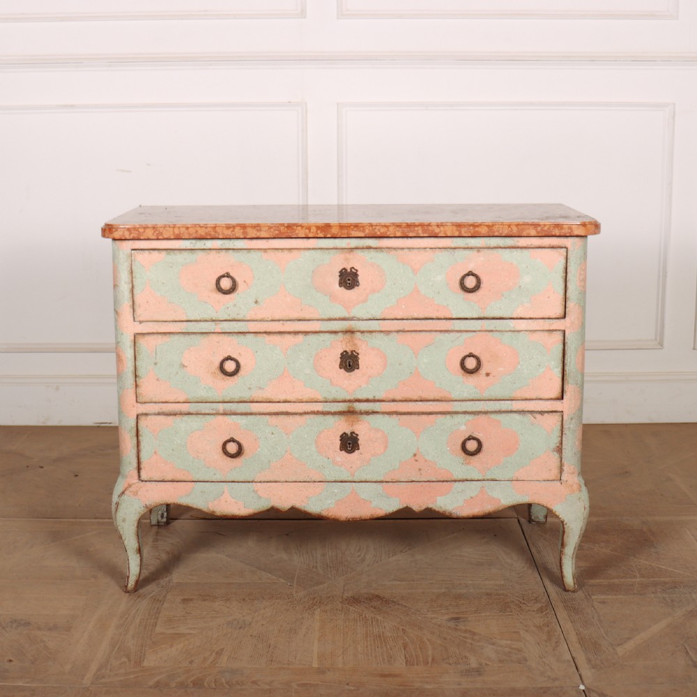 An Italian style three-drawer painted wooden commode featuring a pastel pink and green geometric pattern, brass ring pulls, and a terracotta marble top.