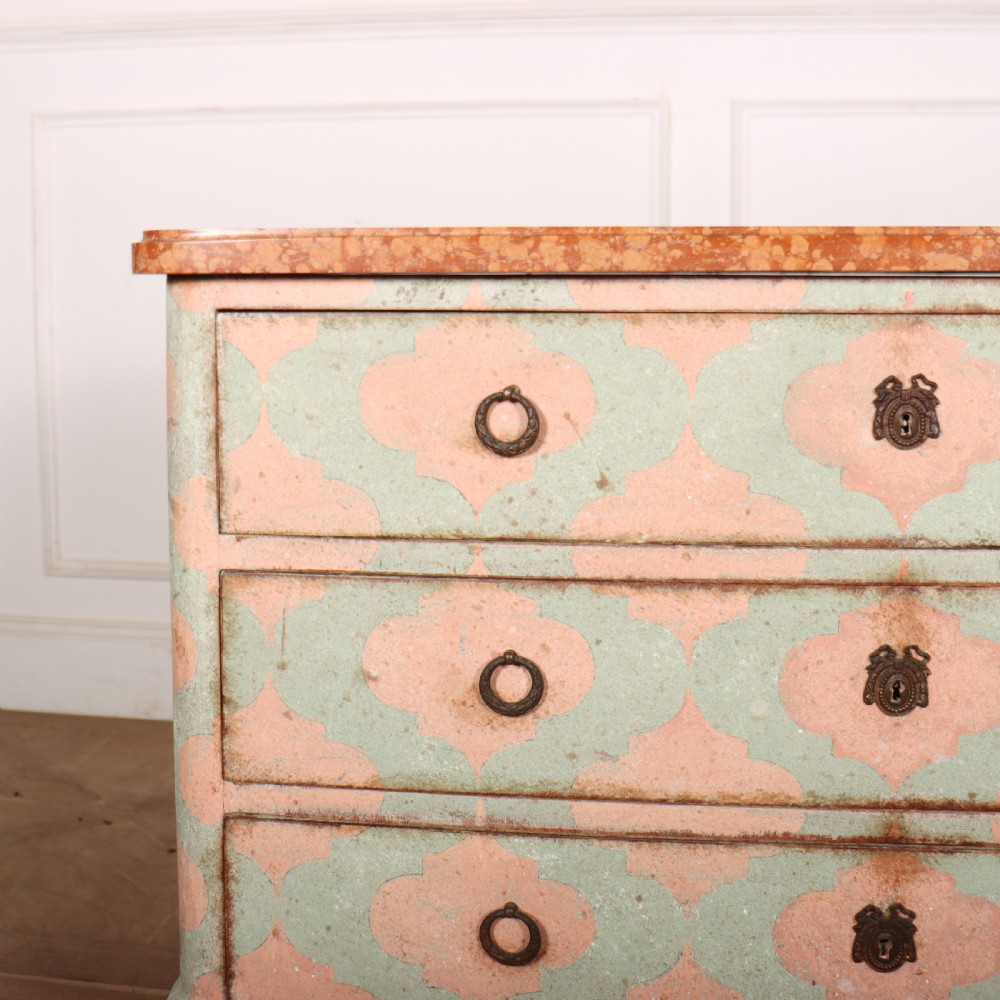 An Italian style three-drawer painted wooden commode featuring a pastel pink and green geometric pattern, brass ring pulls, and a terracotta marble top. View II