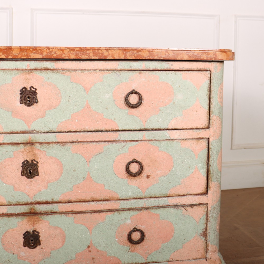 An Italian style three-drawer painted wooden commode featuring a pastel pink and green geometric pattern, brass ring pulls, and a terracotta marble top. View V