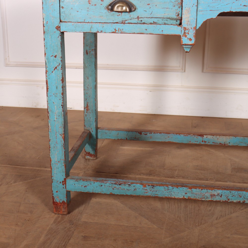 A rectangular wooden table with distressed light blue paint, featuring two drawers with brass cup handles and a low stretcher base. View V