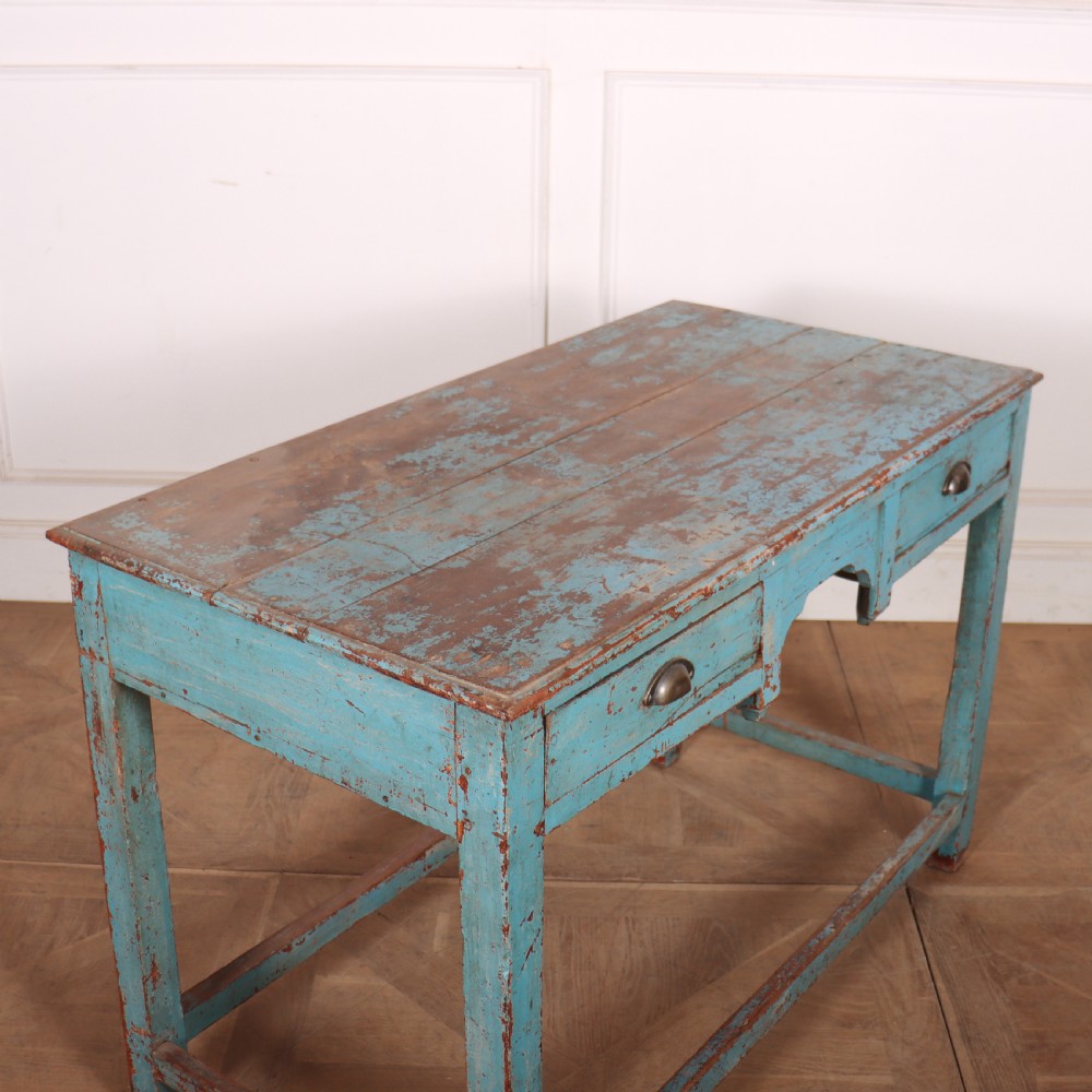 A rectangular wooden table with distressed light blue paint, featuring two drawers with brass cup handles and a low stretcher base. View VI