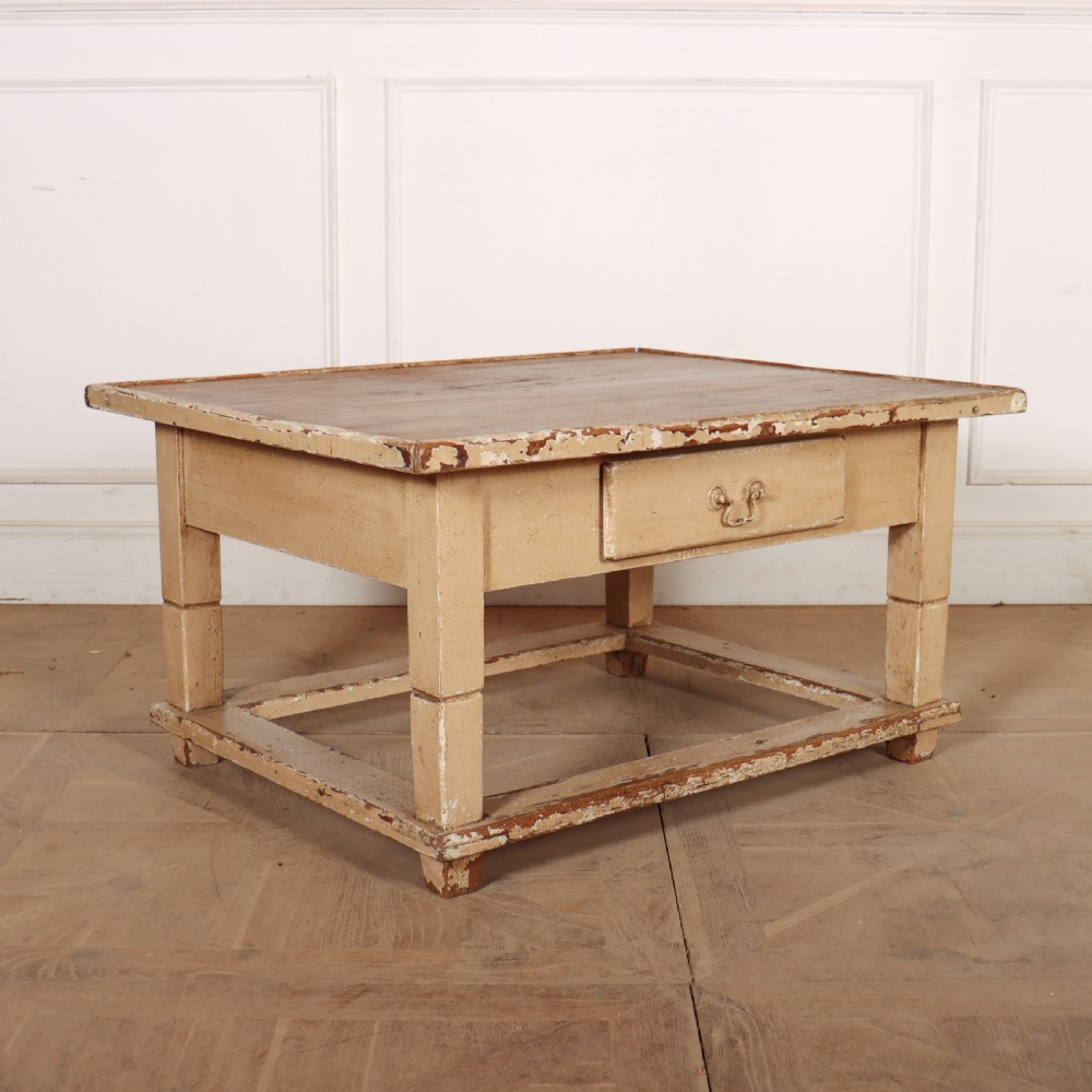A rectangular low painted coffee table featuring a distressed cream paint finish, a single drawer with a metal drop handle, and a perimeter floor stretcher.
